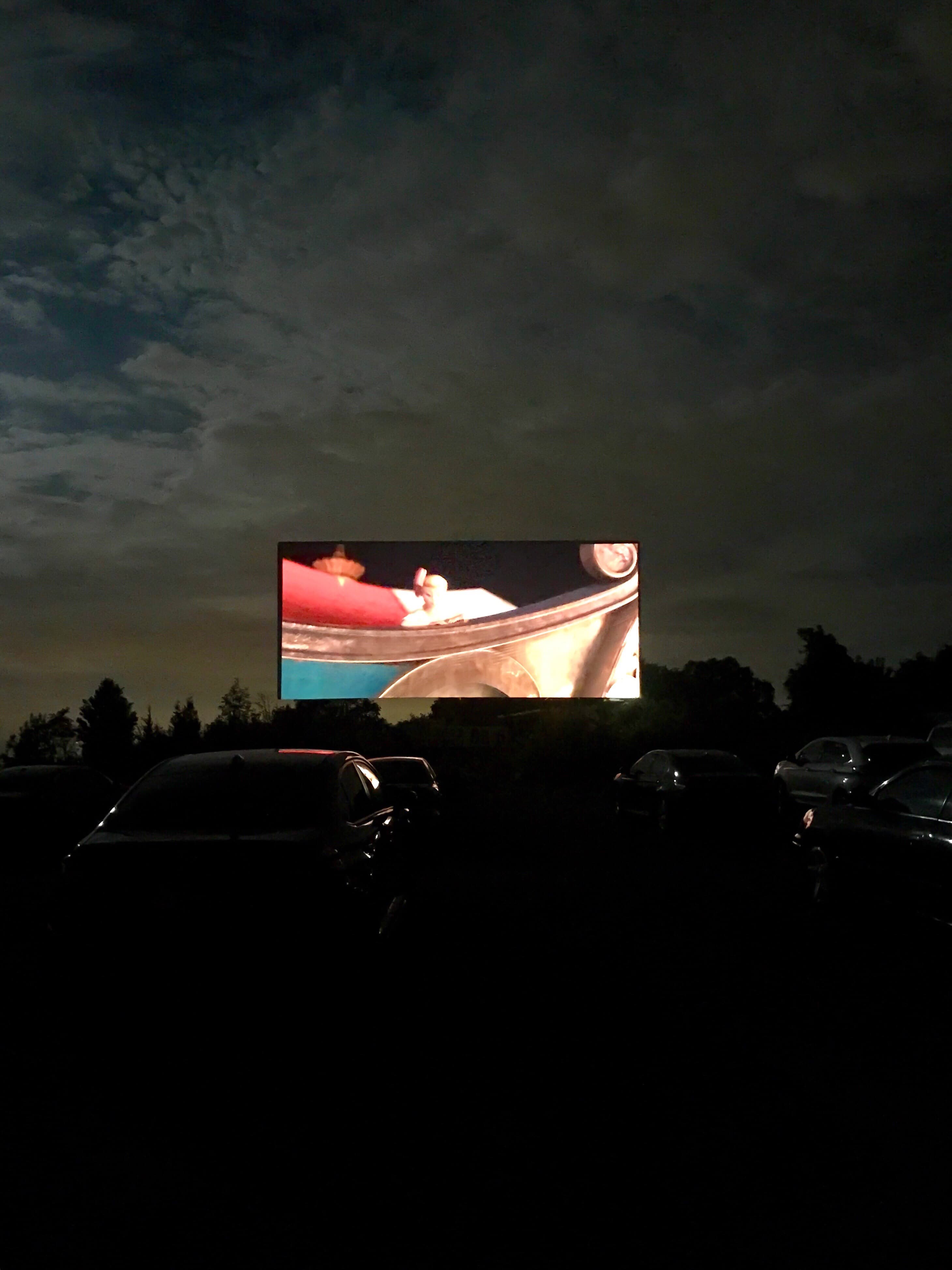 Rows of cars watching a movie on a large outdoor screen at night at a drive-in theater